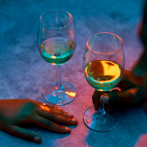 Two people clink glasses of colorful wine in a dim, neon-lit setting, hands resting near the glasses on a table.