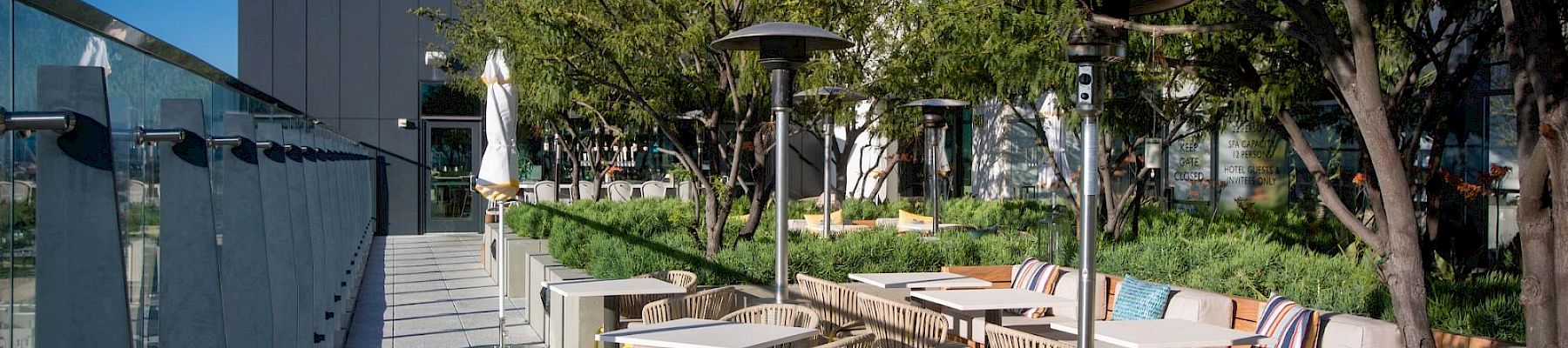 An outdoor cafe area with tables and chairs along a walkway, greenery and trees, modern buildings in the background, sunny sky.