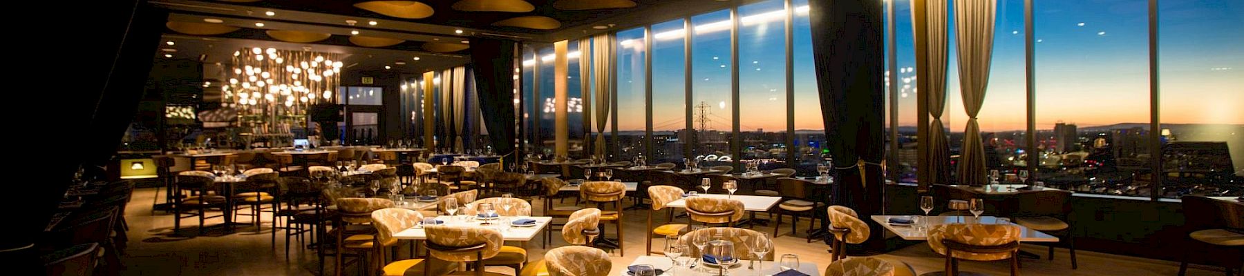 A stylish restaurant with wooden floors, round tables, cushioned chairs, large windows, warm lighting, and a city view at dusk.