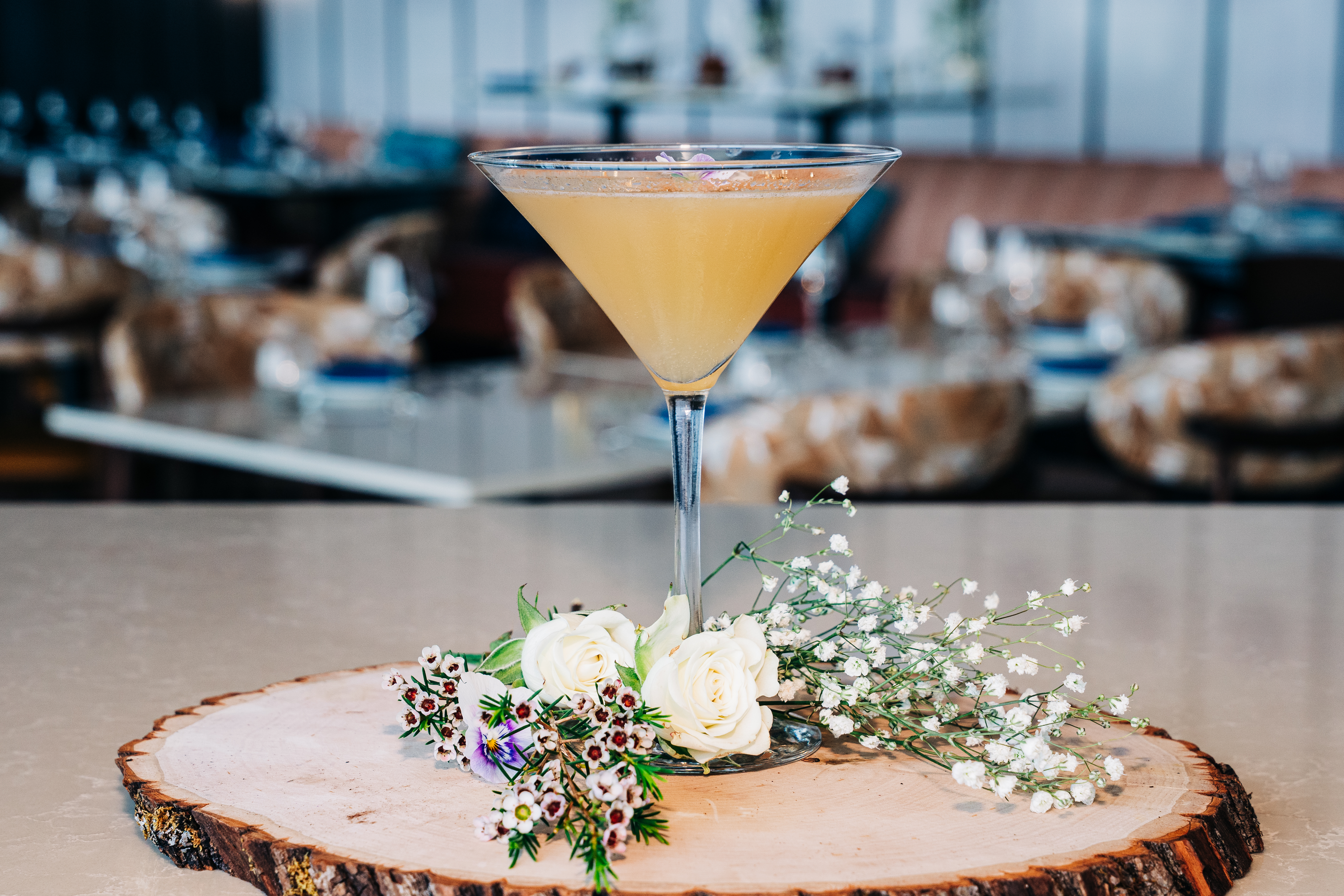 A cocktail in a martini glass sits on a wooden slice centerpiece with small white flowers and greenery, set against a blurred cafe backdrop.