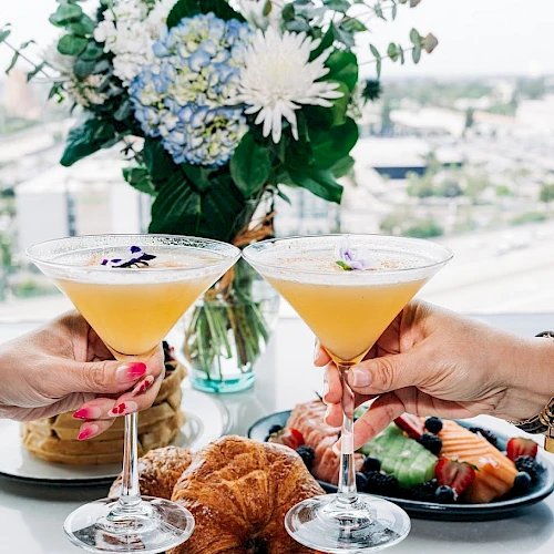 Two people toast cocktails over a chic meal with flowers and pastry bites on a high-rise balcony—celebration vibes in a bright setting.