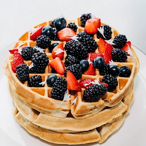 A stack of waffles topped with fresh berries (strawberries, blackberries, blueberries) on a plate, drizzled with syrup. End with period.