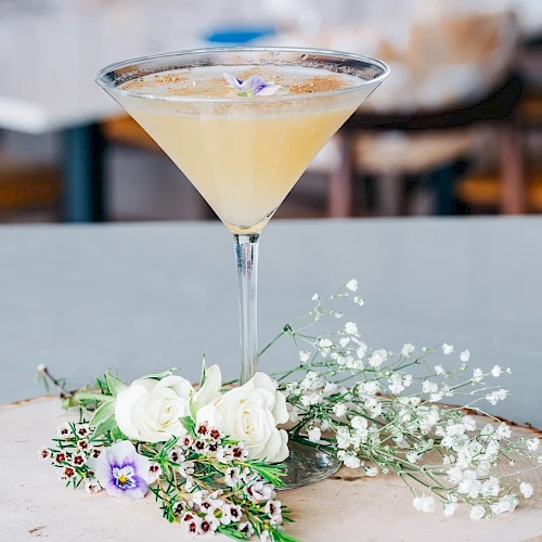 A cocktail in a stemmed glass sits on a wooden slab with a small floral arrangement featuring white blooms and baby's breath.