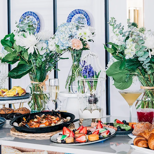 A colorful party spread with pastries, cakes, fruit skewers, and floral centerpieces on a stylish table set against a striped backdrop, festive and inviting.