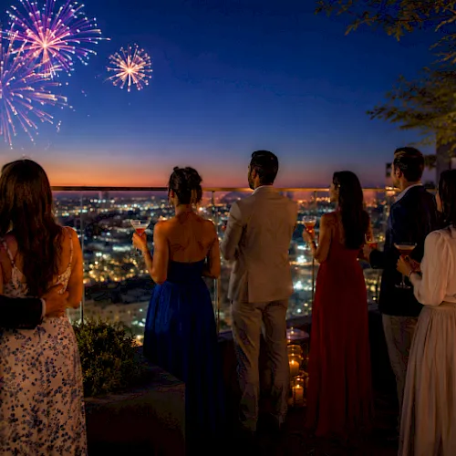 A group of friends on a rooftop overlook watch colorful fireworks explode over a city skyline at dusk.
