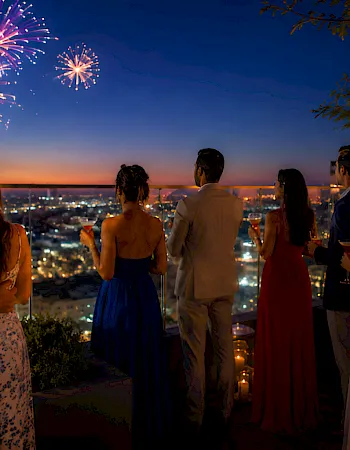 People on a rooftop party watch colorful fireworks over a city skyline at dusk, toasting drinks and celebrating.
