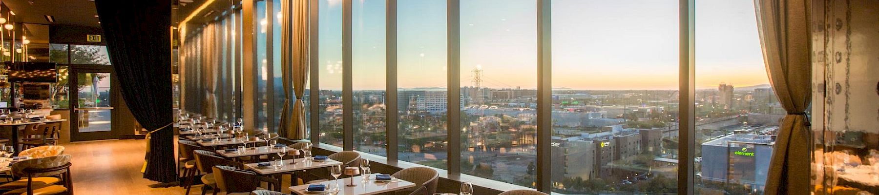 A modern restaurant dining area with set tables, floor-to-ceiling windows, and warm sunset city views in the distance.