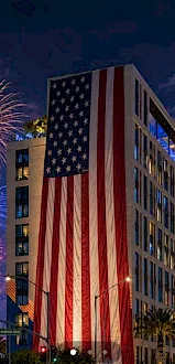 Fireworks light up the sky beside a tall building wrapped in a huge American flag at night.