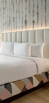 A modern hotel room with a king-size bed, soft curtains, lamps, and a cozy seating area by the window, with warm lighting at dusk.