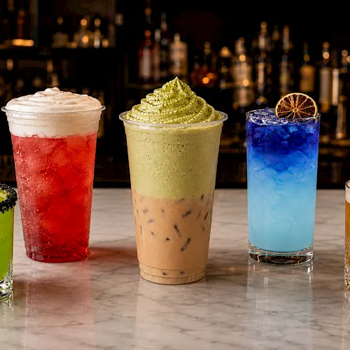 Four colorful cocktails—green, red, pale yellow, and blue—plus a golden amber drink with citrus garnish, on a bar counter.