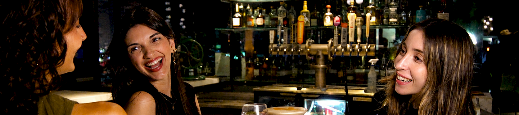Three friends clink glasses and smile at a bar, celebrating with drinks under warm lights.