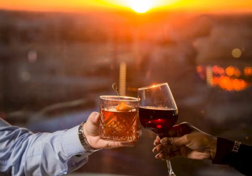 Two people toast with cocktails over a sunset dinner&mdash;glasses raised above bowls of food as the warm sky glows in orange hues.