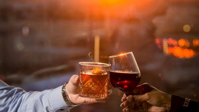 Two people toast with cocktails over a sunset dinner&mdash;glasses raised above bowls of food as the warm sky glows in orange hues.