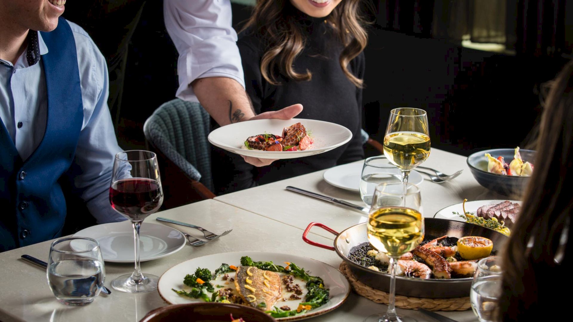 A group of people at a table enjoying a fancy dinner with wine, plates of food, and a server presenting a dish, in a lively dining scene.