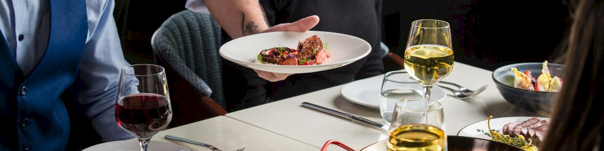 A group of people at a table enjoying a fancy dinner with wine, plates of food, and a server presenting a dish, in a lively dining scene.