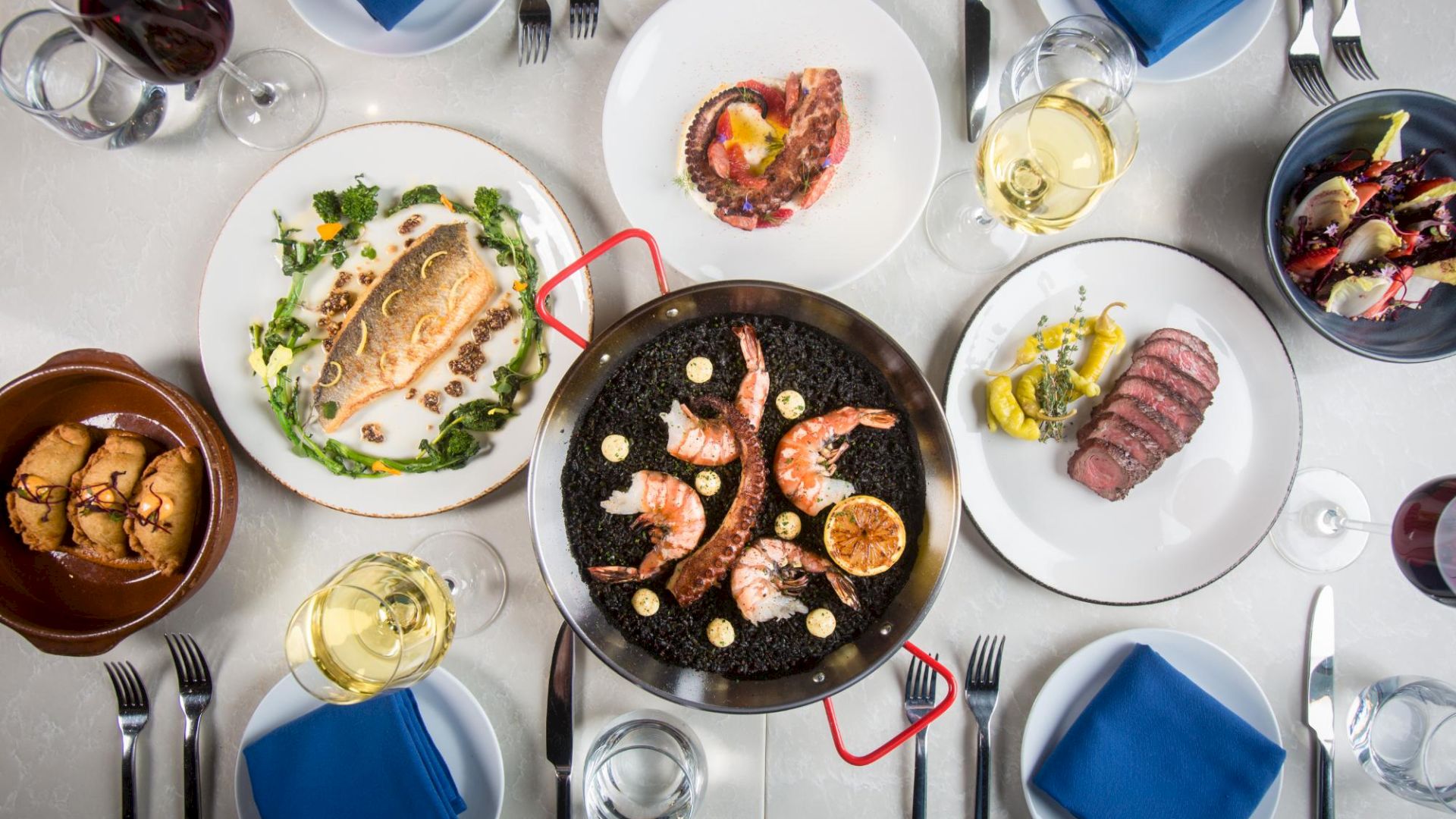 A top-down view of a table loaded with seafood and meats: shrimp on a grill pan, breaded items, sliced steak, pasta, and assorted sides, with wine glasses.