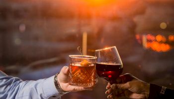 Two people toast with drinks at sunset over a table with bowls and plates of food, creating a warm, romantic dinner scene.