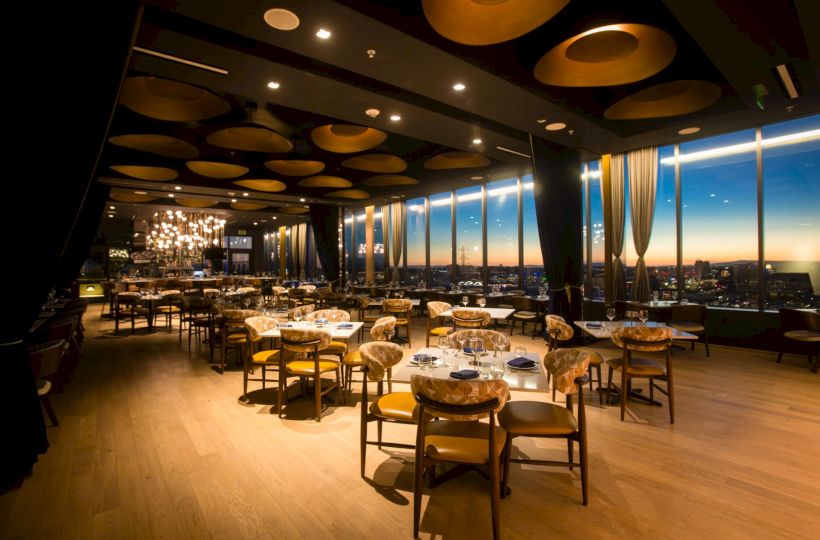 A stylish dining room restaurant with wooden floors, round tables, chairs, large windows, and warm lighting over sunset views, elegant and upscale.