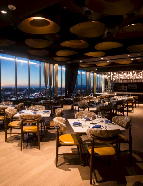 A stylish restaurant with large windows, city views at dusk, round tables set with plates and glassware, yellow chairs, and warm ambient lighting.