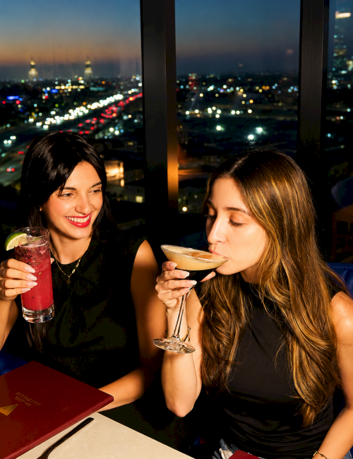 Two women smile and sip cocktails at a nighttime rooftop, with city lights glowing beyond the window.