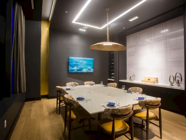 A modern private dining room with a long white table, plush yellow chairs, a wall-mounted screen, and contemporary lighting in a stylish, dark-toned space.