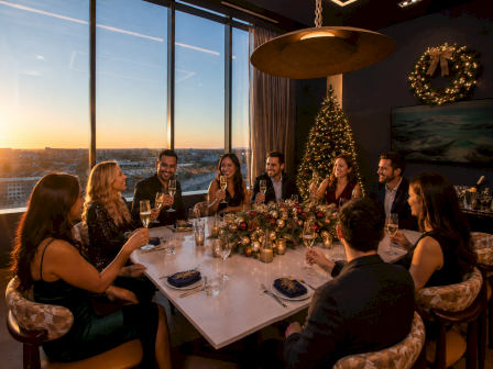 A group of friends cheers with champagne at a festive holiday dinner by a window, lit by a decorated Christmas tree.