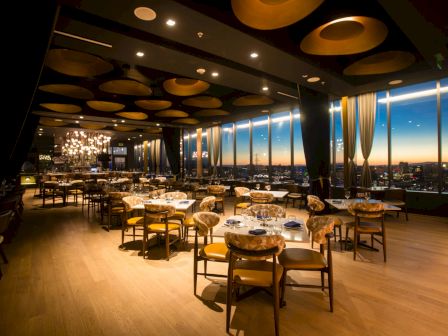 A modern fine-dining restaurant with set tables, warm chandeliers, and tall windows overlooking a sunset city skyline.