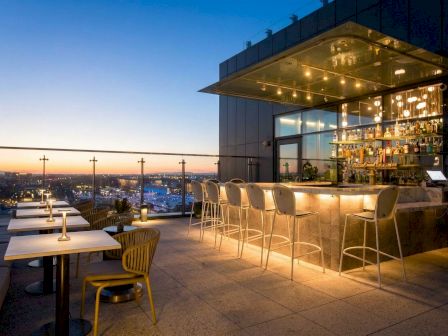 A modern rooftop bar at dusk with a glass canopy, glowing liquor bottles, stools, and a city skyline view beyond.
