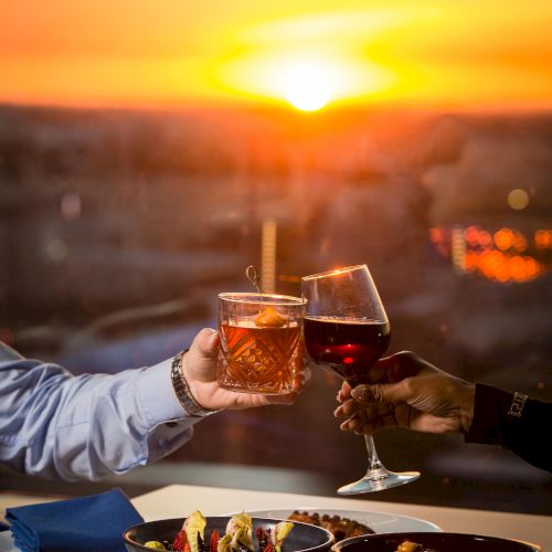 A sunset dinner scene with friends toasting drinks over plates of food on a balcony.