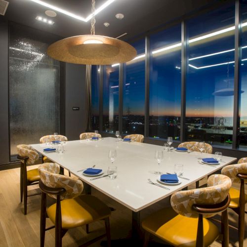 A modern office lounge with a long wooden table, pendant lights, and a city view through large windows at night.