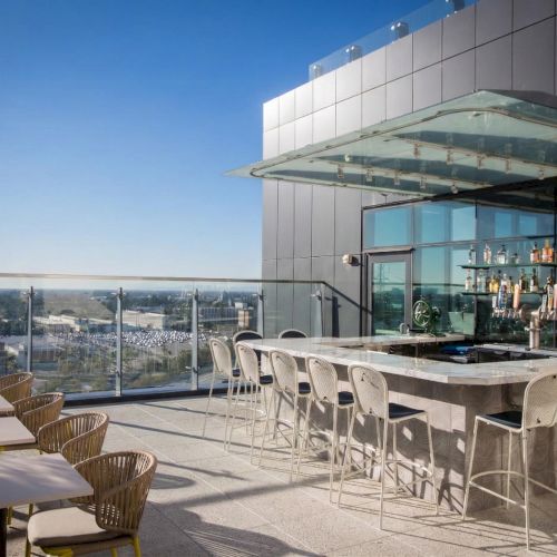 A rooftop bar with a long marble counter, white bar stools, wicker chairs, and glass railing overlooking a cityscape on a sunny day.