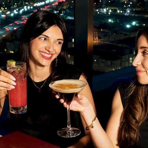 Two women toast at a rooftop bar with colorful cocktails as the city lights blur in the night background.