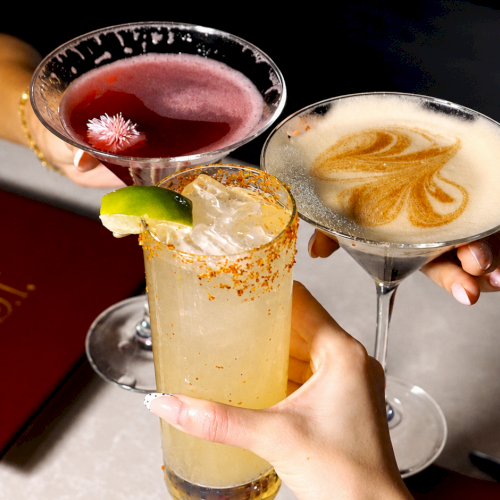 Hands clink cocktails&mdash;one red with a flower, one creamy orange swirl&mdash;beside a frothy golden drink on a tray.