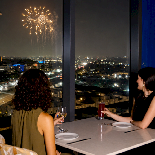 Two people dine by a window watching fireworks over a city at night, with drinks on the table.