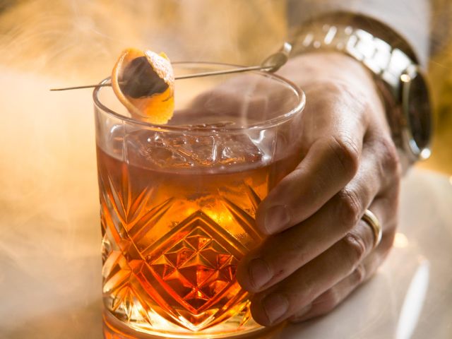 A hand holds an amber cocktail with ice cubes and an orange peel garnish in a cut-glass tumbler.