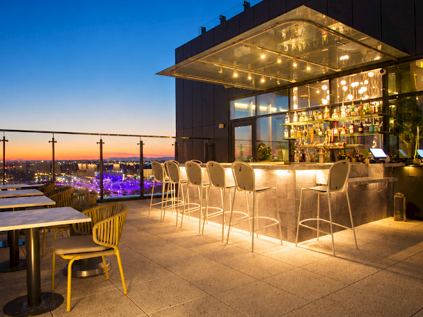 A rooftop bar terrace with lit bottles and white stools, overlooking a city skyline at dusk under a deep blue sky.