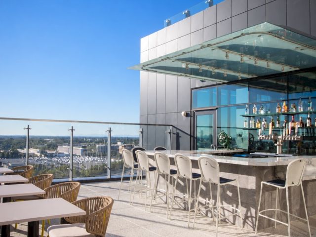 A modern rooftop bar patio with white chairs, a glass-topped counter stocked with bottles, and a view under a clear blue sky.