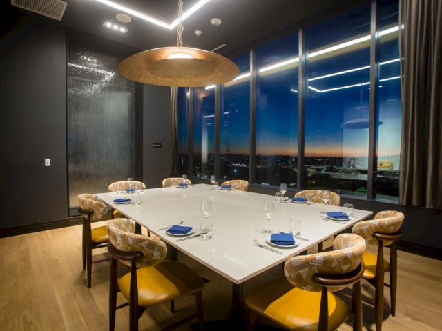 A modern dining room with a set table, upholstered chairs, a gold chandelier, and big windows showing the city skyline at dusk.