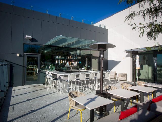 A sunny rooftop patio with outdoor seating, heaters, and a glass-fronted bar counter between modern building walls.