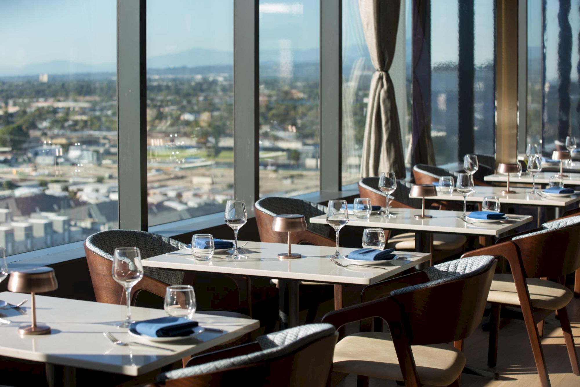 A stylish restaurant with big windows, neatly set tables, wine glasses, and wooden chairs overlooking a cityscape; bright, modern, inviting.