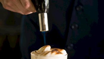A bartender pours a shot from a bottle into a glass of frothy whiskey, crafting a layered, foamy cocktail on a marble bar.