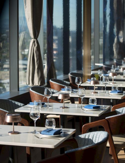 A stylish restaurant with a sunlit dining area, white tables, blue napkins, wine glasses, and wooden chairs along a glass wall with curtains.
