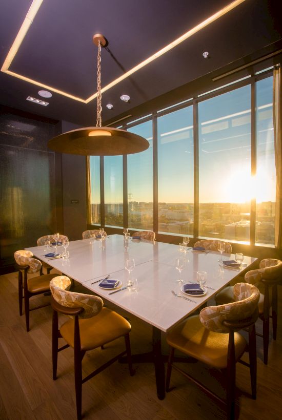 A stylish dining room with a long white table, eight cushioned chairs, large windows overlooking a sunset, and a hanging pendant light above the table.