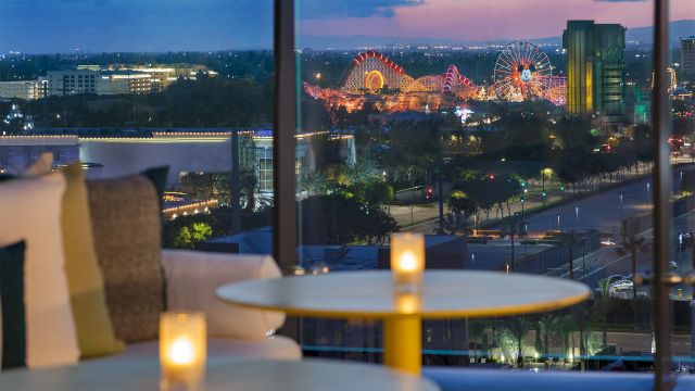 A cozy indoor lounge with candles on a round table, glass windows, and a nighttime cityscape with a lit Ferris wheel outside. Top it at 140 characters, always ending the sentence.