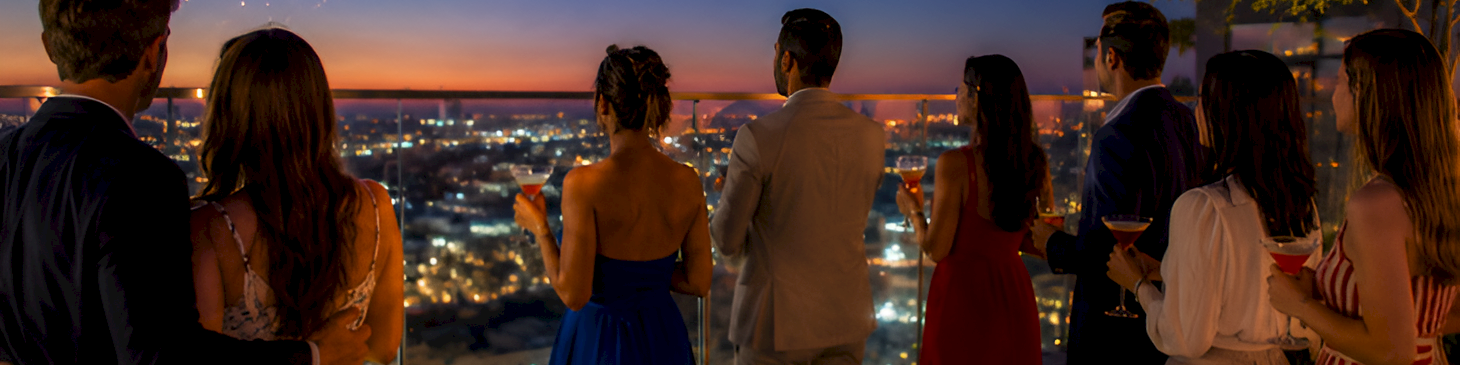 A rooftop party crowd watches colorful fireworks over a city skyline at dusk.