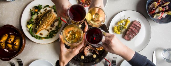 People toast with assorted dishes and drinks around a table, sharing a meal that includes steak, salad, bread, and colorful beverages.