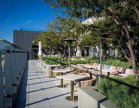 An outdoor cafe area with tables and chairs along a walkway, greenery and trees, modern buildings in the background, sunny sky.