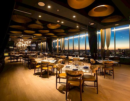 A stylish restaurant with wooden floors, round tables, cushioned chairs, large windows, warm lighting, and a city view at dusk.