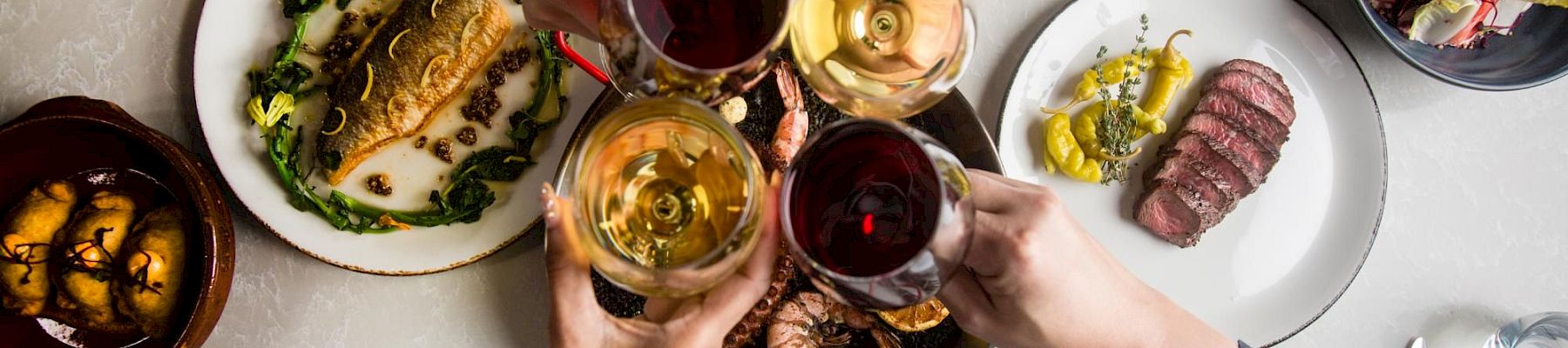 A group toasts with wine glasses over a table of diverse dishes, including steaks, greens, and sides, sharing a celebratory meal.
