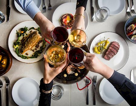 A group toasts with wine glasses over a table of diverse dishes, including steaks, greens, and sides, sharing a celebratory meal.